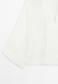 Biała krótka koszula z szerokimi rękawami, wykonana z lekkiego materiału. Posiada dekolt w kształcie litery V oraz proste linie, co podkreśla minimalistyczny design.
