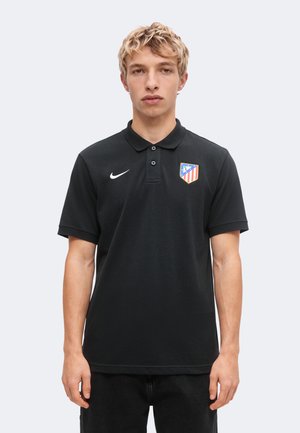 Młody mężczyzna z krótkimi blond włosami, ubrany w czarną koszulkę polo Nike z logo klubu piłkarskiego Atletico Madryt, stoi na jednolitym jasnym tle.