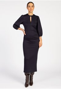 Frau in einem schwarzen Midi-Kleid mit drei Viertel Puffärmeln und Schlüsselausschnitt, die braune kniehohe Stiefel trägt und vor einem weißen Hintergrund steht.