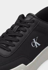 Μαύρο χαμηλό αθλητικό παπούτσι με μαύρα κορδόνια, λευκό λογότυπο CK στο πλάι και λευκή σόλα, με πάνω μέρος από λείο συνθετικό υλικό.