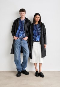 Two models wear coordinated outfits. The male model has a black hooded coat and blue patterned sweater, paired with light jeans and black shoes. The female model wears a similar coat over a blue patterned sweater and white skirt, with black footwear.
