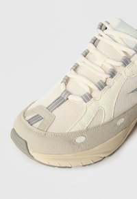 Bēša sporta apavu pāris ar acs un zamšādas materiāliem, pelēkiem akcentiem, teksturētu zoli un plakanām auklām, radīts komfortam un stilam.