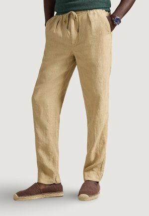 Hombre con pantalones de lino beige con cordón, alpargatas marrones sin cordones, una camiseta verde y un reloj de pulsera, con las manos en los bolsillos.