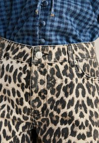 Jean en denim léopard avec une base beige et des taches noires, doté d'un bouton en métal argenté et de coutures détaillées sur les poches.