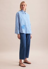 Camicia a righe blu e bianche con design a bottoni, caratterizzata da un ricamo blu. Abbinata a pantaloni in denim blu larghi e sandali neri.