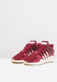 Červené pletené tenisky s bílými pruhy, mají strukturovaný svršek, gumovou podrážku a šněrování. Zdobení zahrnuje polstrovaný límec a jazyk.