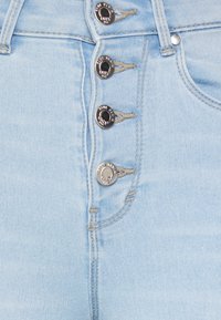 Jeans de denim azul claro apresentando quatro fechos de botão com detalhes metálicos, costura contrastante e uma textura suave no tecido.