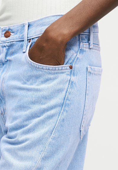 Tume nahaga käsi on pandud valgelt sinise teksapükste esimesse taskusse, mida kannab valge pluus.