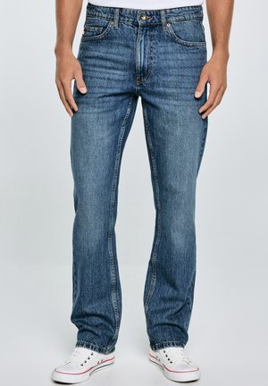 Vaqueros de denim azul con corte de pierna recta, costuras prominentes y un diseño clásico de cinco bolsillos, combinados con zapatillas de lona blancas.
