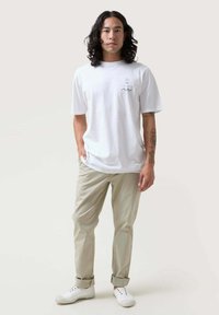 Camiseta blanca de algodón con un pequeño diseño gráfico, combinada con chinos beige claros y zapatillas blancas, mostradas en un modelo masculino.