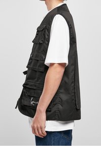 Černá vesta s několika kapsami, vyrobená z odolné tkaniny. Má zapínání na zip, druky a na přední straně je charakteristické logo.