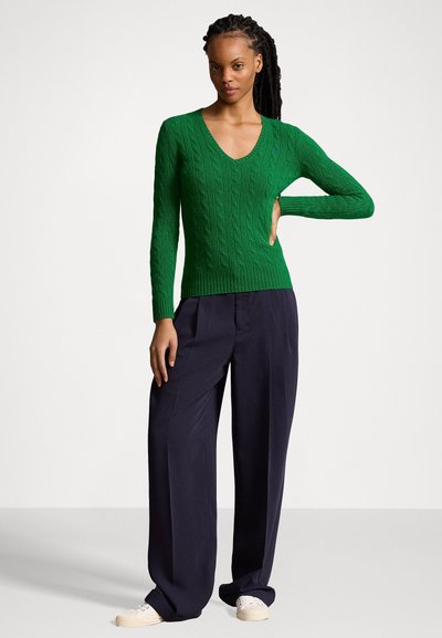 Zelený svetr s pleteným vzorem a výstřihem do V, úzký v pase, kombinovaný s volnými tmavými kalhotami a světlými botami.