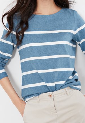 Femme portant une chemise bleue à manches longues avec des rayures blanches horizontales rentrée dans un pantalon beige, avec une main dans la poche.