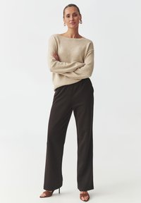 Beige gebreide trui met een relaxte pasvorm, gecombineerd met zwarte wijde broek. Het model draagt bruine haksandalen en grote hoepeloorringen.
