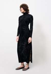 Schwarzes Samtkleid mit floralem Muster, hohem Kragen, langen Ärmeln und Seitenschlitzen. Getragen mit schwarzen Netzballerinas und Knöchelriemen.