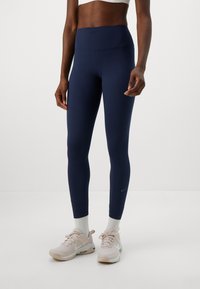 Person med marinblå högmidjade leggings, vita strumpor och ljusrosa träningssneakers som står mot en enkel vit bakgrund.