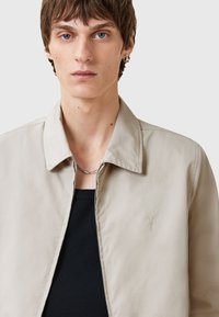 Beige lichtgewicht jas met een gladde textuur, voorzien van een spitse kraag, rits aan de voorkant en subtiele logo borduursel aan de linkerkant.