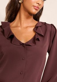 Brūna blūze ar viļņotu apkakli, pogu priekšējo aizdari un gludu tekstūru. Tuvplāns uz apkakles un augšējām auduma detaļām.