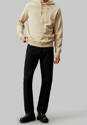 Felpa beige in cotone con cappuccio e stringhe, con dettaglio del logo; abbinata a jeans neri dritti in denim e scarpe nere stringate.