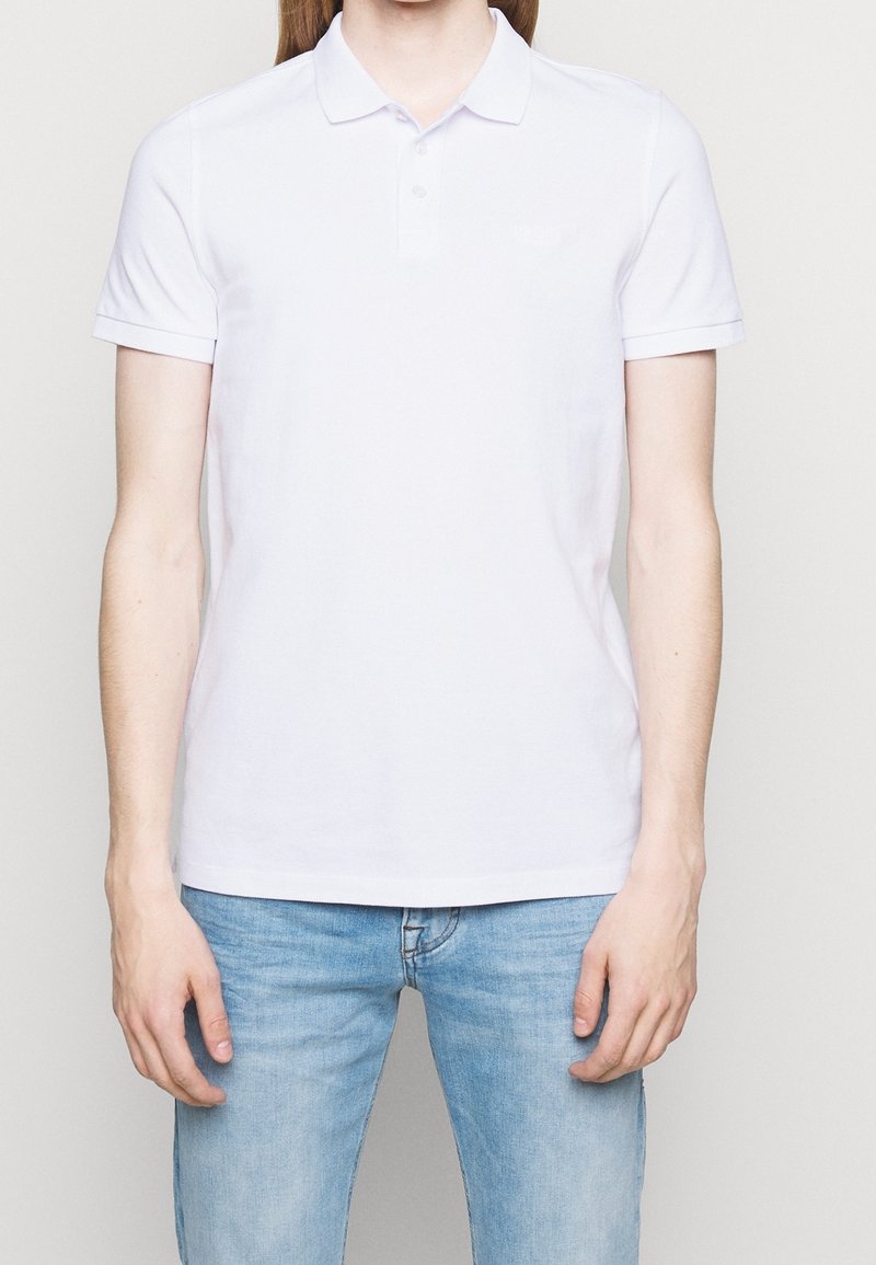Polo en coton blanc à manches courtes, avec col et trois boutons sur la patte de boutonnage. Texture lisse, design simple, sans motifs apparents.