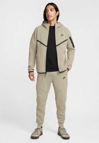 Nike vetoketjullinen huppari ja jogginghousut vaaleanbeigessä, joissa on kontrastiväriset mustat yksityiskohdat, vetoketjulliset taskut ja sileä, teksturoitu pinta.