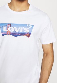 Valkoinen puuvillapaita, jossa on suuri Levi's-logo ja kuva Golden Gate -sillasta kirkkaissa väreissä sinistä taustaa vasten.