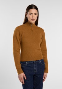 Brown zip-neck sweater in soft knit fabric with ribbed cuffs and hem, paired with dark denim jeans. Structured, fitted design.
