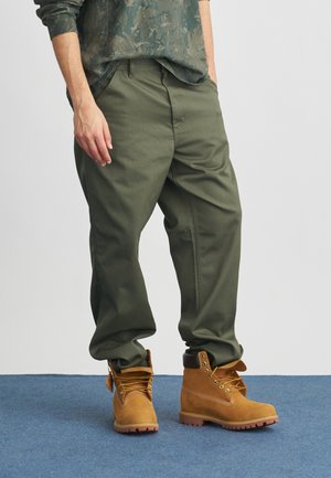 Pantalones de algodón verde combinados con botas de cuero marrón. Los pantalones tienen un corte holgado y las botas cuentan con cordones dorados y una suela resistente.