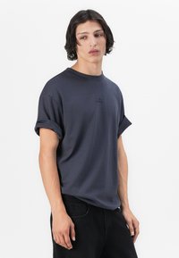 Czarnoszary t-shirt z okrągłym dekoltem, wykonany z miękkiej bawełny. Posiada podwinięte rękawy i małe logo na klatce piersiowej. Luźny krój.