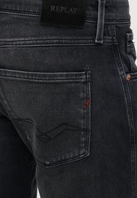 Κοντινή εικόνα μαύρου τζιν από denim που δείχνει την πίσω τσέπη με διακριτικό ραφτό σχέδιο, μικρή κόκκινη λεπτομέρεια "R" και μαύρη ετικέτα Replay στη μέση.