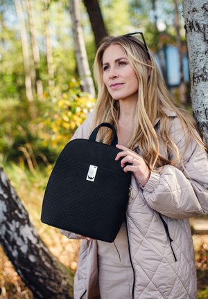 Černý texturovaný batoh s prošívaným designem, se stříbrným logem a horním madlem, kterého se drží osoba v lehké kůži venku.