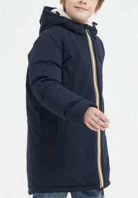 Veste matelassée bleu marine avec une capuche doublée de fausse fourrure, détail de zip jaune et poches latérales. Le tissu semble lisse avec du volume et de la chaleur.