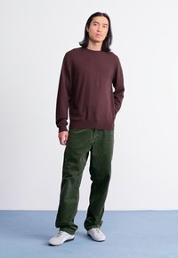 Suéter de punto marrón con cuello redondo, combinado con pantalones de pana verdes y zapatillas blancas, de pie sobre un suelo azul contra un fondo blanco.