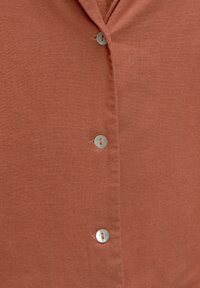 Chemise boutonnée en terracotta avec une texture lisse, dotée d'un col à encoche et de boutons en métal argenté sur le devant.