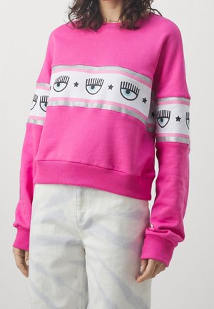 Personne portant un sweat-shirt rose vif avec des bandes blanches affichant des yeux bleus et des étoiles noires sur la poitrine et les manches, accompagné d’un pantalon clair.