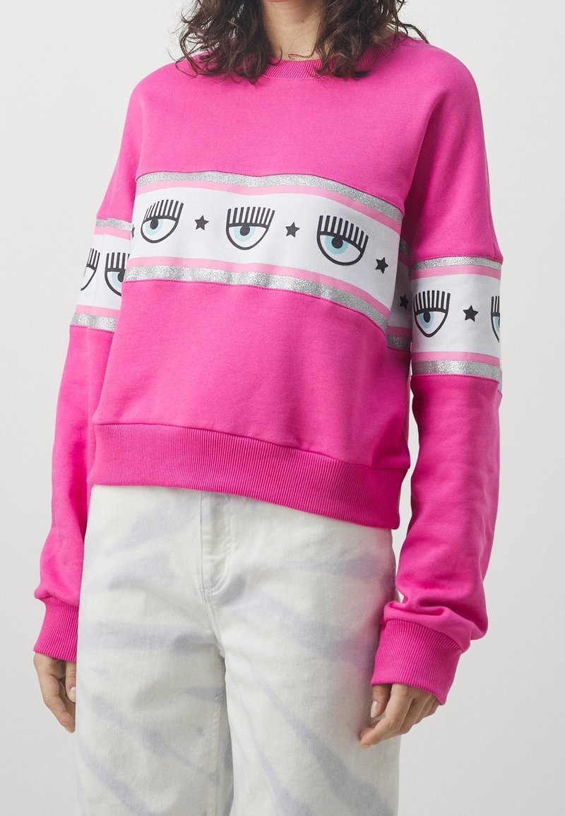 Personne portant un sweat-shirt rose vif avec des bandes blanches affichant des yeux bleus et des étoiles noires sur la poitrine et les manches, accompagné d’un pantalon clair.