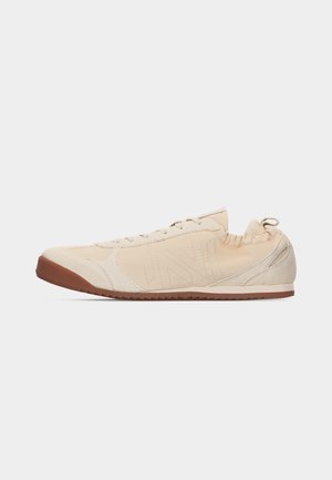 Beiger Sneaker mit niedrigem Profil, Wildledereinsätzen, Schnürverschluss und brauner Gummisohle, seitlich auf hellem Hintergrund dargestellt.