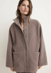 Veste en polaire de couleur marron clair, au design ample avec un col haut, de grandes poches latérales et une fermeture à boutons à l'avant. Texture douce et apparence confortable.