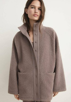 Chaqueta de forro polar en marrón claro, diseño cuadrado con cuello alto, grandes bolsillos laterales y cierre frontal de botones. Textura suave y apariencia acogedora.