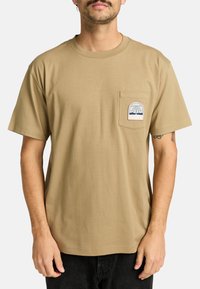 Beige katoenen t-shirt met een ronde hals, korte mouwen en een linkerkant borstzak met een palmboomgrafiek en RVCA-logo.