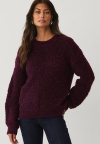 Vrouw met lang bruin haar, die zilveren oorringen draagt, een gestructureerde donkerpaarse trui en donkerblauwe spijkerbroek, staande tegen een effen achtergrond.