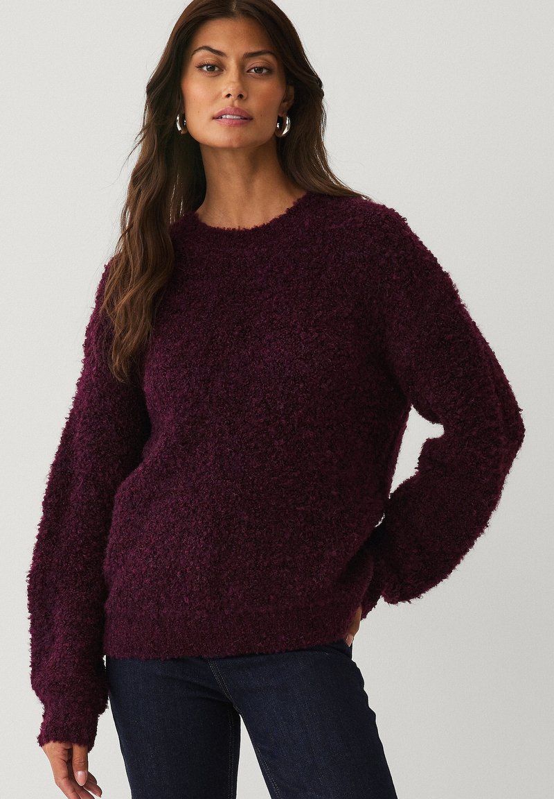 Kvinde med langt brunt hår iført sølv hoops-øreringe, en struktureret mørk lilla sweater og mørkeblå jeans, der står foran en ensfarvet baggrund.
