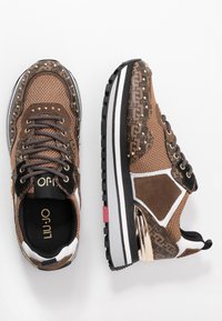 Zapatillas marrones de malla y ante con detalles metálicos, tachuelas y un panel lateral estampado. Cuentan con una suela blanca gruesa con dibujo negro.