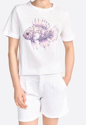Femme portant une chemise blanche à manches courtes et un short, la chemise présente un grand motif de poisson composé d'ornements en forme de bijoux violets et transparents.