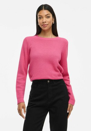 Pull en maille texturée rose avec des manches longues et un col rond, associé à un jean noir taille haute. Le style est ajusté et décontracté.