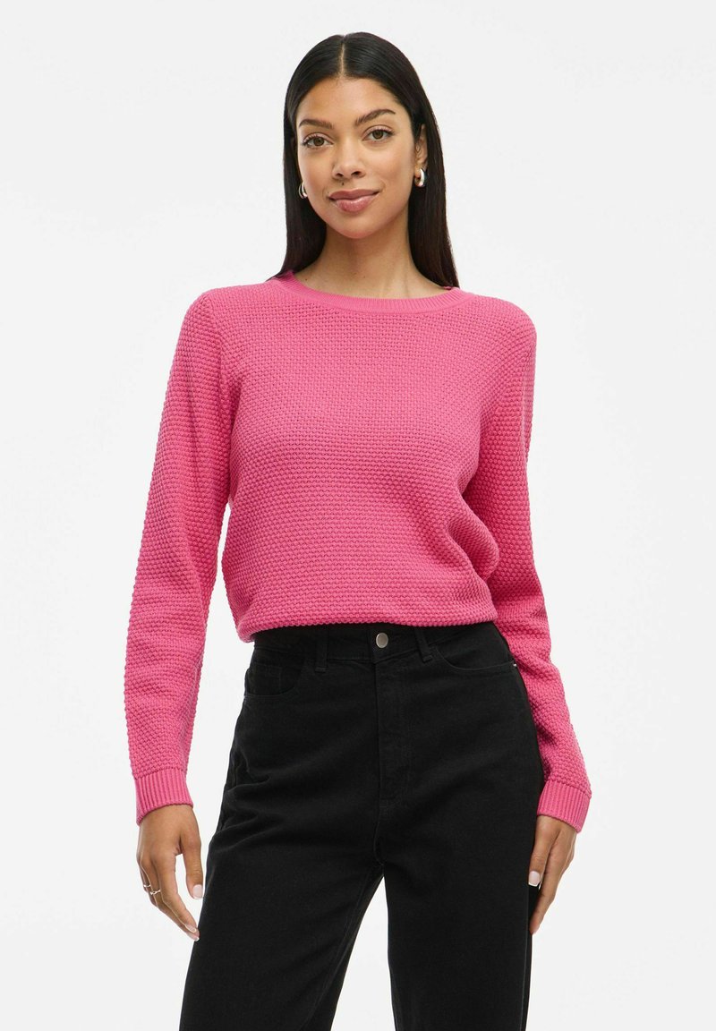 Roze gestructureerde gebreide trui met lange mouwen en een ronde halslijn, gecombineerd met hoge zwarte jeans. De stijl is aansluitend en casual.