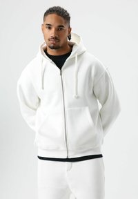 Sweat à capuche blanc zippé avec une coupe décontractée, cordons, poches avant et une texture douce, assorti à un pantalon blanc et un t-shirt noir.