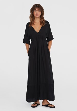 Robe maxi noire avec des manches courtes en aile de chauve-souris, décolleté en V, taille froncée, poches sur les côtés et un ourlet à volants fluide. Stylée avec des tongs.