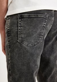 Close-up van een persoon die vervaagde zwarte spijkerbroek draagt, waarbij de achterzak en een deel van een wit overhemd zichtbaar zijn.