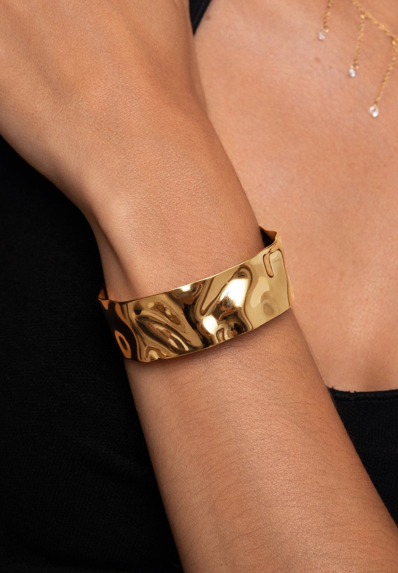 Bracciale rigido in oro con una texture liscia e ondulata, caratterizzato da un design aperto e una finitura lucida. Indossato su un polso contro un tessuto scuro.